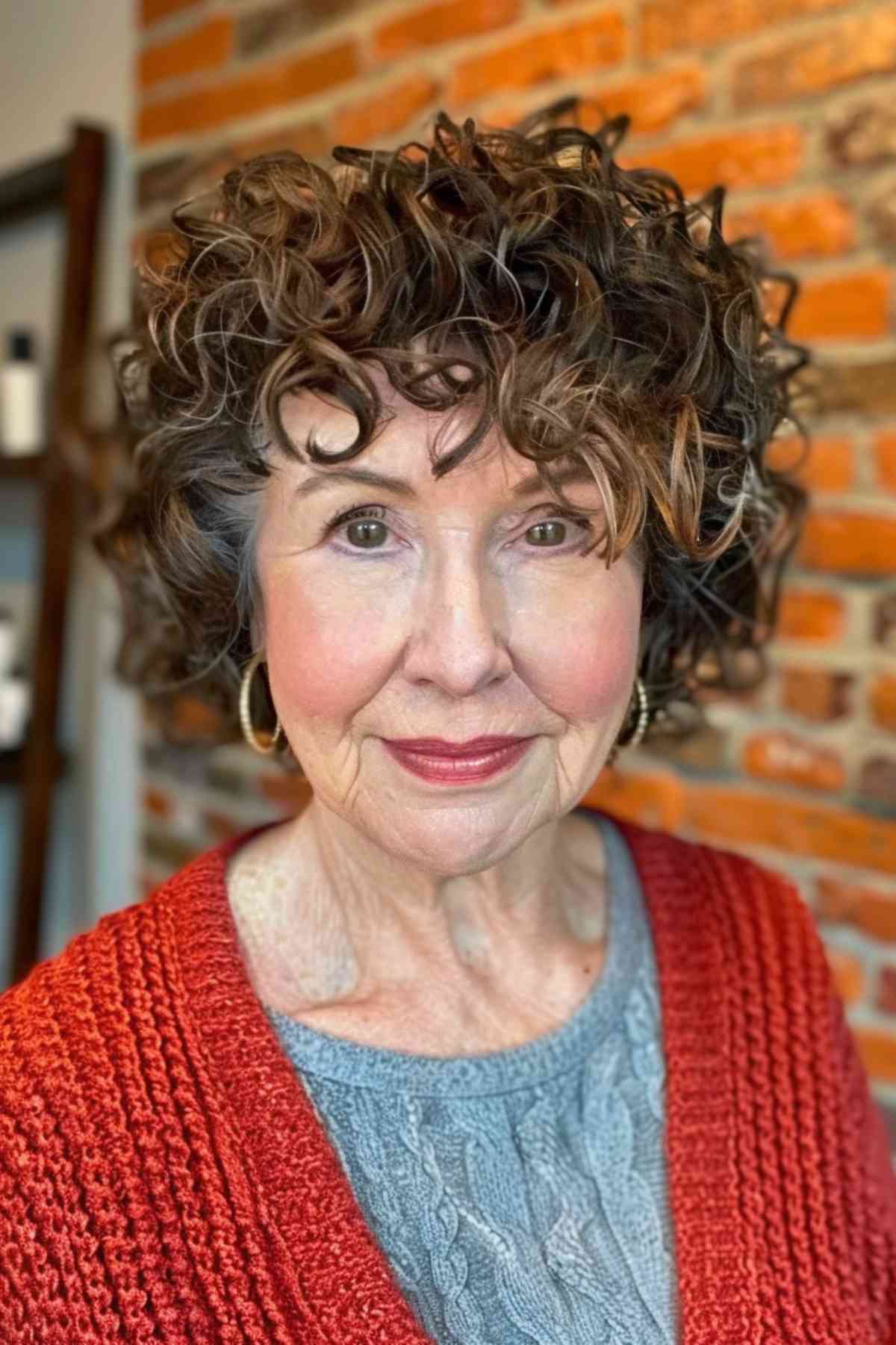 Mature woman with natural curly hair and soft bangs, wearing a red knitted sweater and blue underlay.